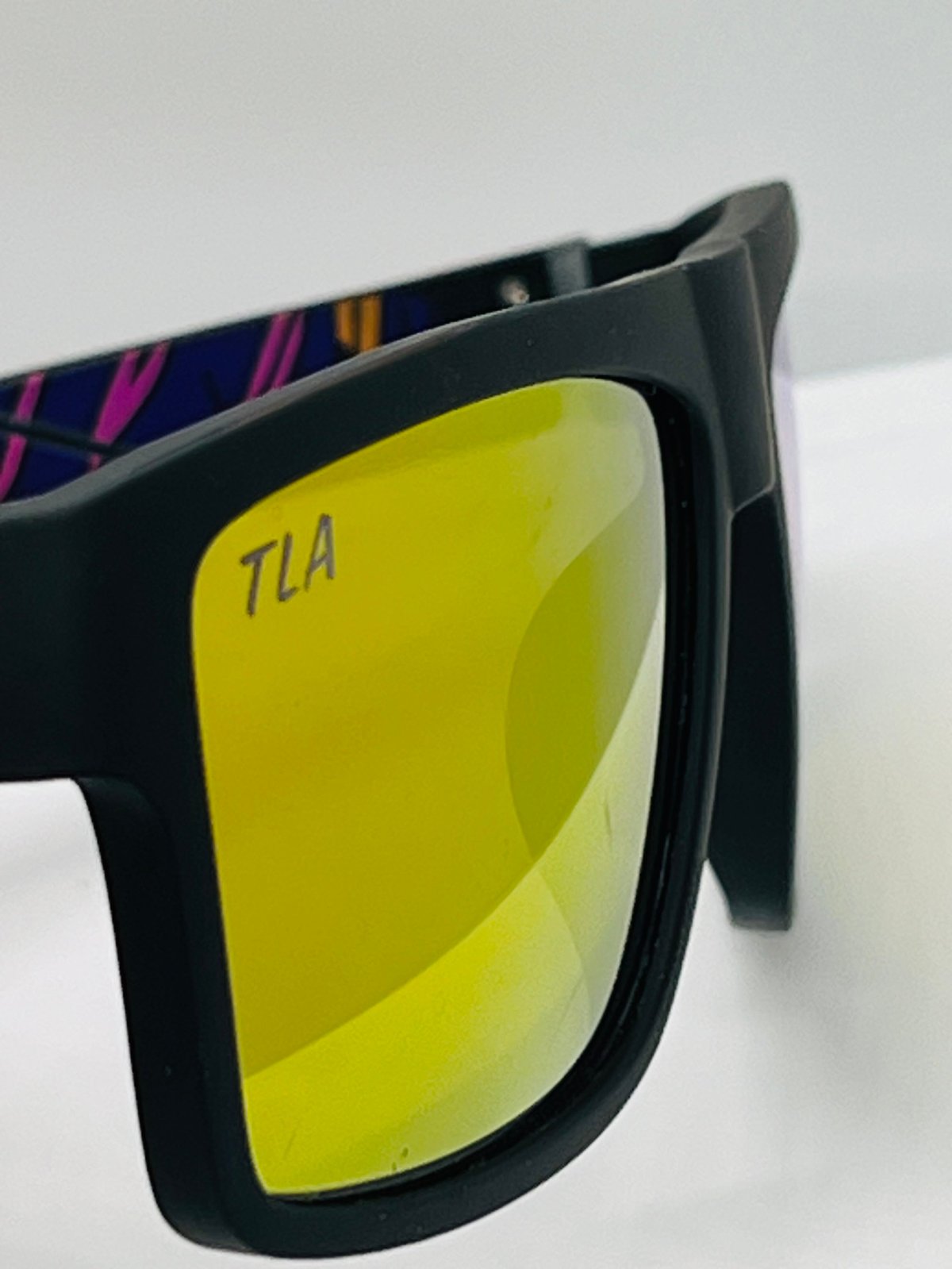 Black-framed sunglasses with TLA text etched on the yellow mirrored lens and colorful striped arms