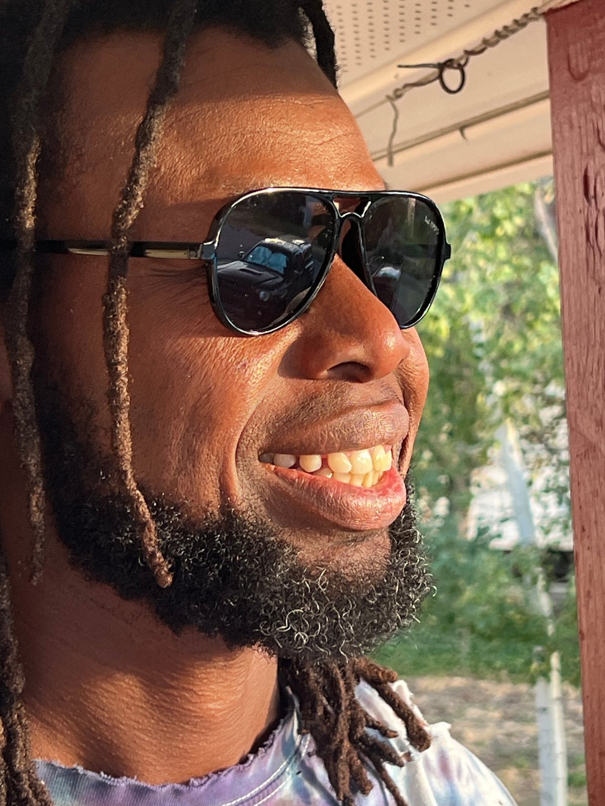 Man with braided hair and beard wearing dark aviator sunglasses, smiling outdoors in daylight