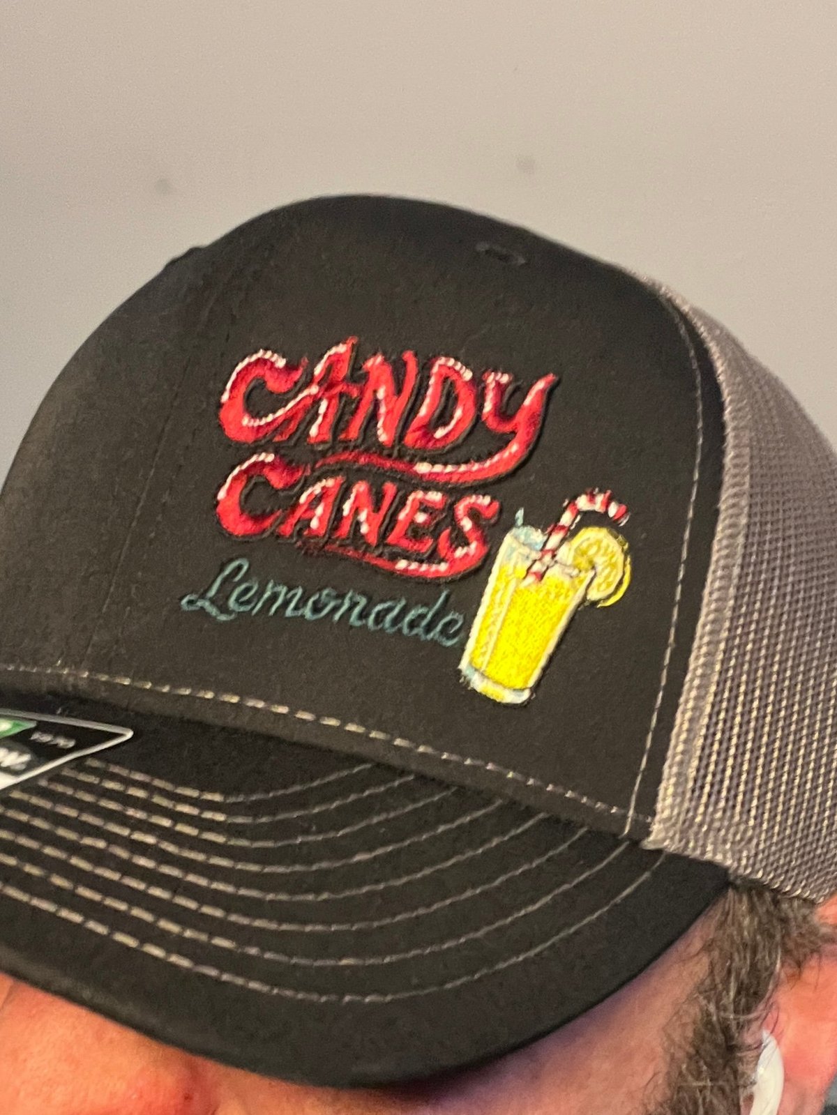 Black and gray trucker cap with red Candy Canes text and yellow lemonade glass embroidery, worn on person with visible shoulder