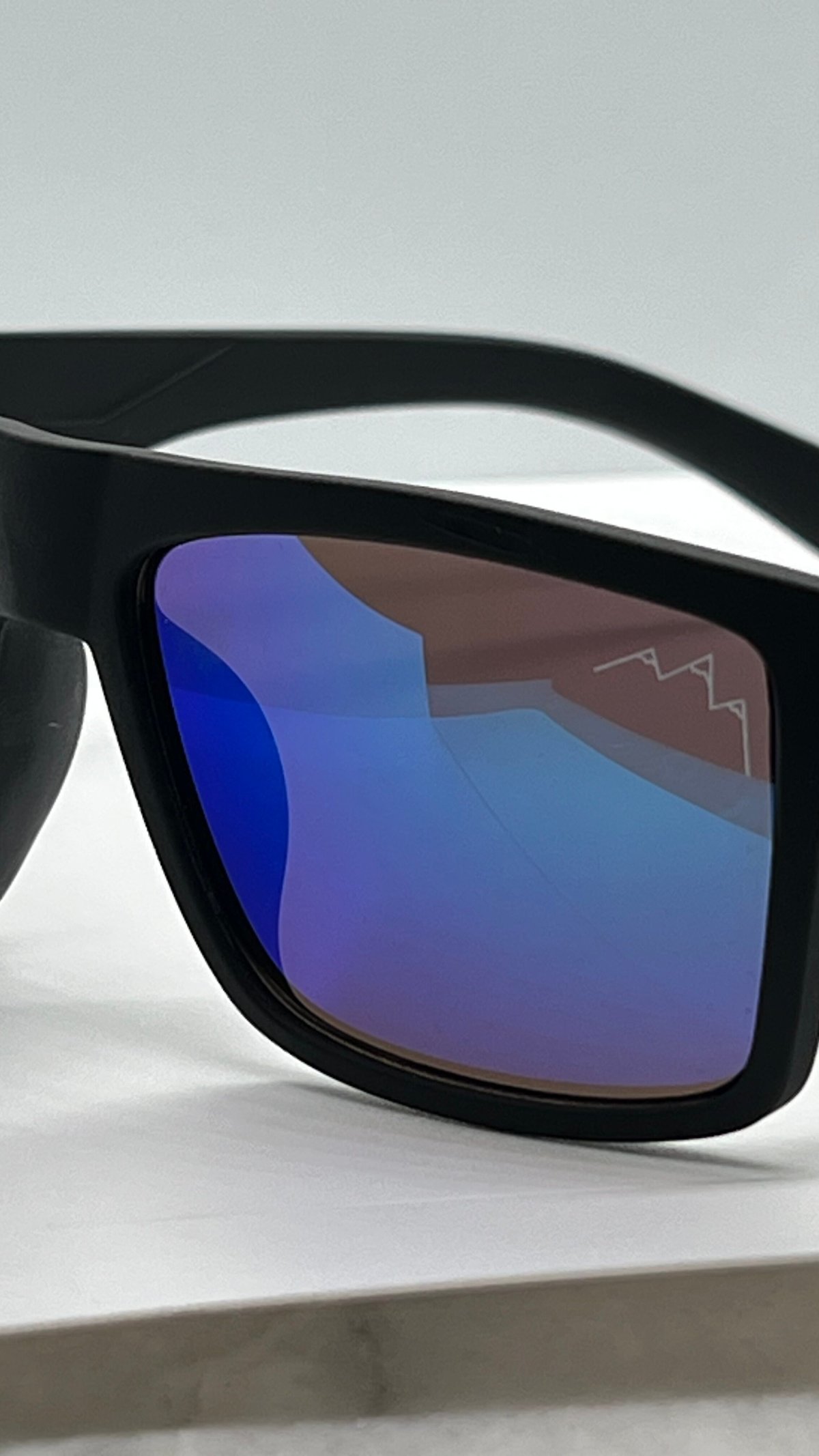 Black-framed sunglasses with the TLA mountain logo etched on the blue gradient mirrored lens