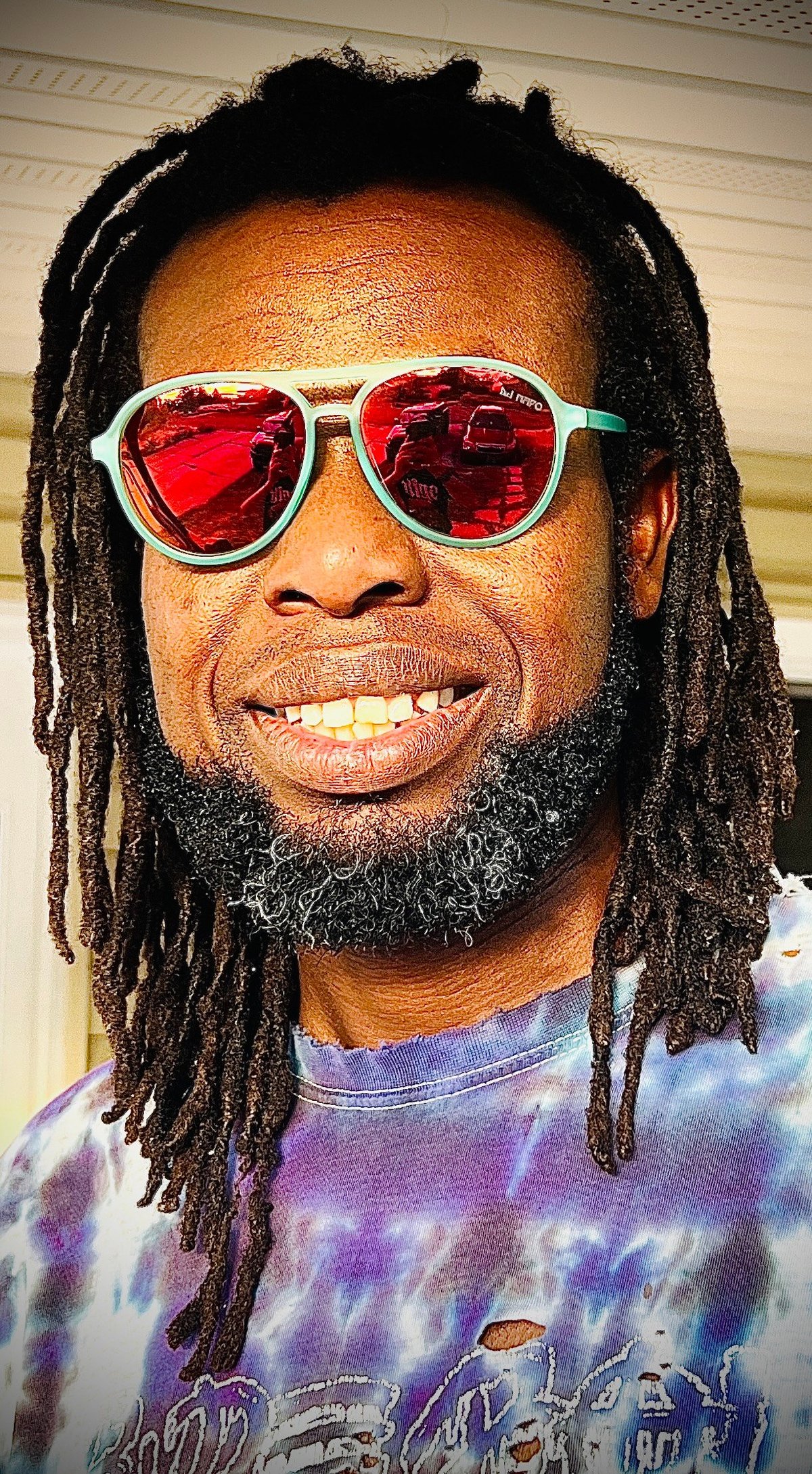 Man with dreadlocks wearing white-framed heart-shaped red sunglasses and a tie-dye shirt, smiling at camera