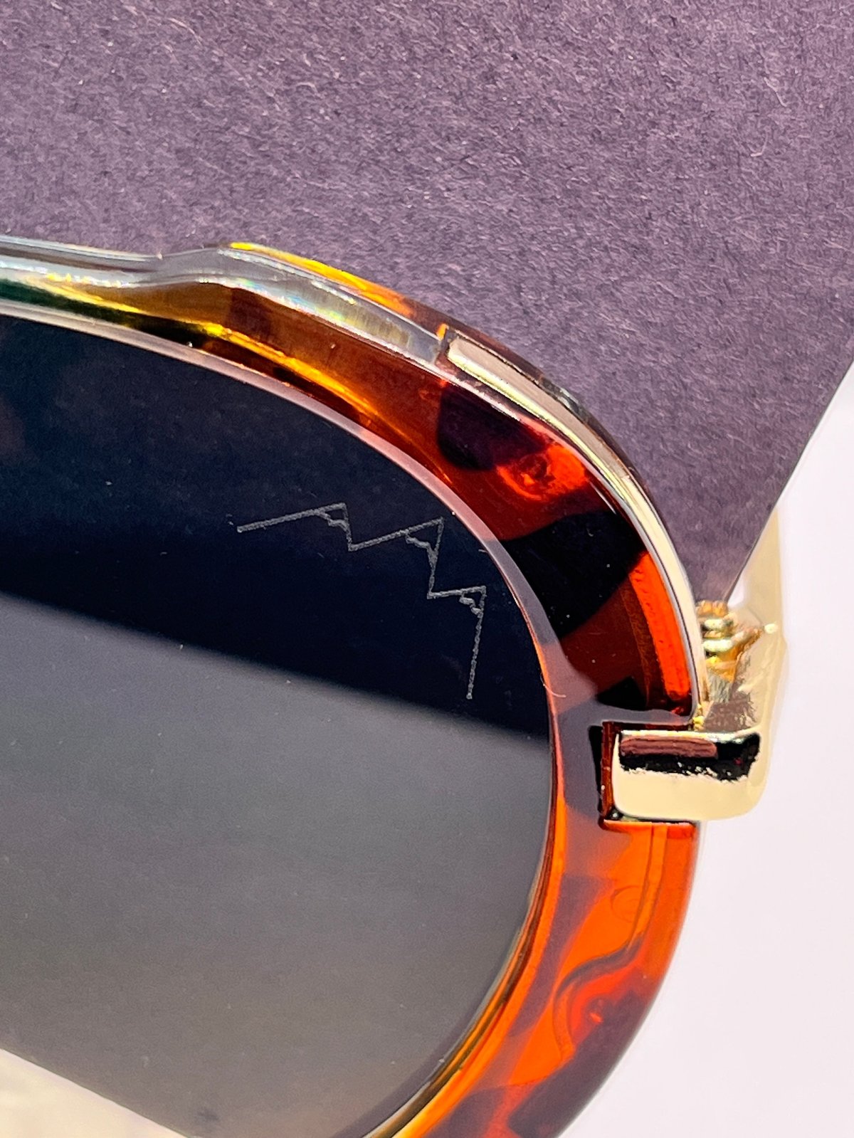 Gold Digger Teal Tortoise sunglasses with amber tortoiseshell frames, the TLA mountain logo etched on the lens against a purple background