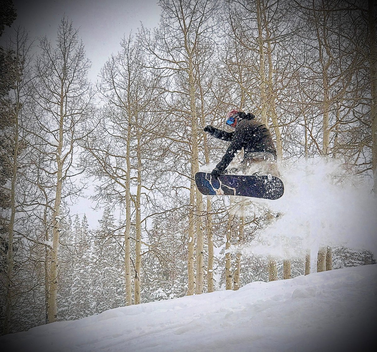 Ben snowboarding, performing a jump in mid-air on a snowy slope with bare trees in the background
